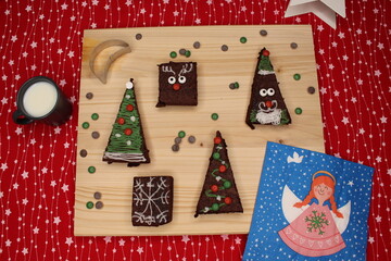 Five brownies in the shape of a Christmas tree, Santa Claus and reindeer. On top of a wooden table. There are chocolate drops and a glass of milk. In the background, a red tablecloth with white stars.