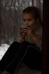 A child with a cup of tea near a winter window with a blurred background.