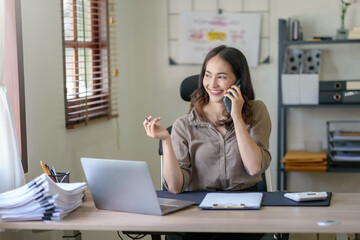 Asian businesswoman sitting happily working on a laptop and talk on the phone to contact customers, talk to explain the job and smile happily.