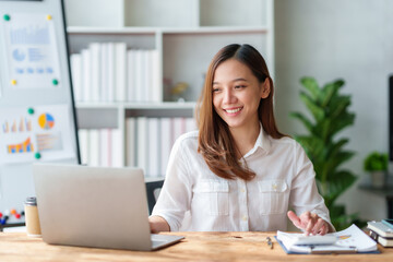 Pretty and cute Asian businesswoman smiling and enjoying coming up with ideas for working with laptop and document.