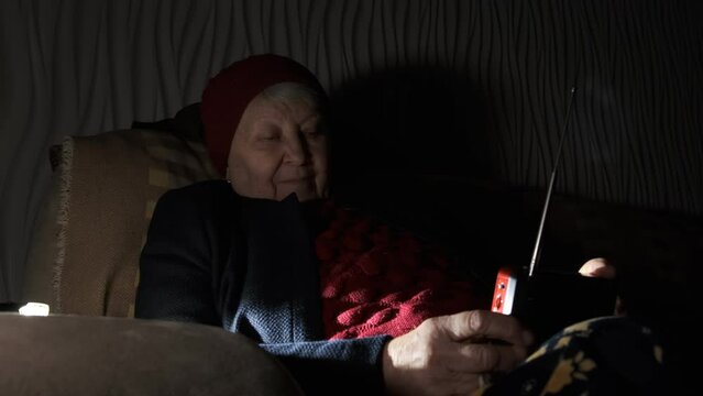 Grandmother holds radio in hands sitting in darkness blackout in Ukraine during russian terroristic attacks. Senior woman in winter clothes in darkness on sofa with hand radio receiver listening news.