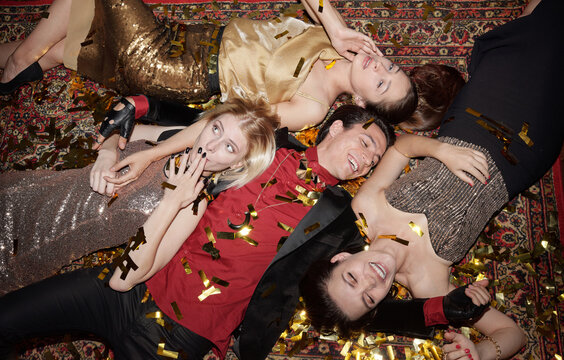 Four boozy friends having fun posing on camera lying on floor in confetti at party