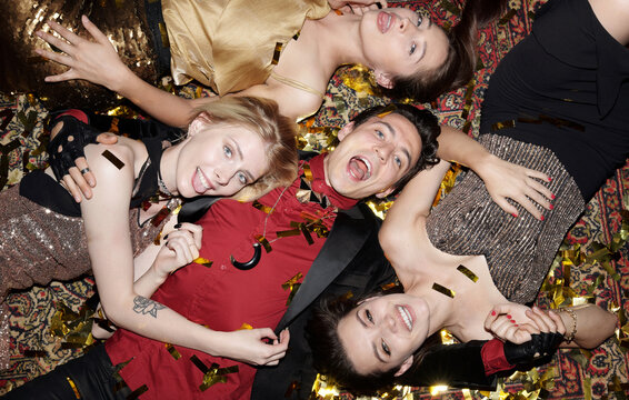 Four models wearing fashionable shiny clothes lying together on rug grimacing on camera