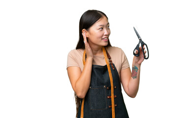 Young Asian seamstress woman over isolated background listening to something by putting hand on the ear