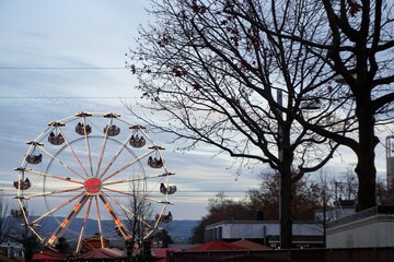Bäume vor Dächern von Hüttendächern auf Weihnachtsmarkt und Riesenrad vor Himmel am Nachmittag...
