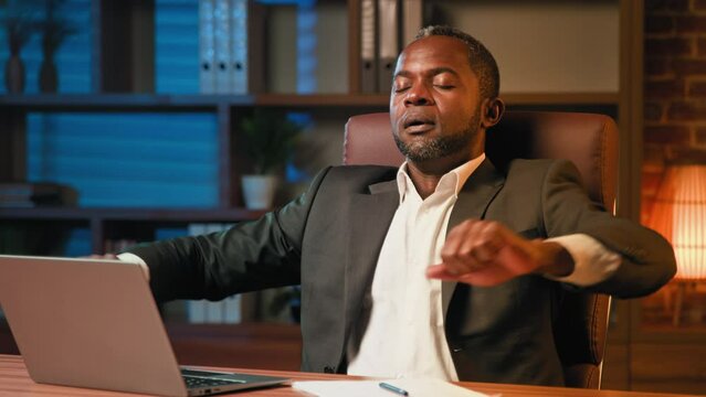Focused adult businessman working until late in office using laptop tired man finished computer work stretching sitting at workplace desk feeling relieved after job well done putting hands behind head