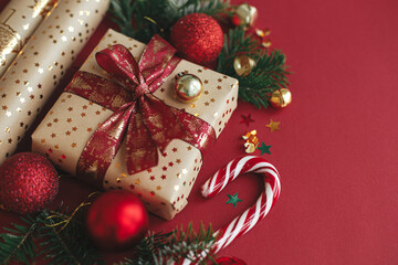 Modern christmas composition. Stylish christmas gift, wrapping paper,  fir branches, candy cane and baubles on red background. Merry Christmas and Happy Holidays! Festive banner