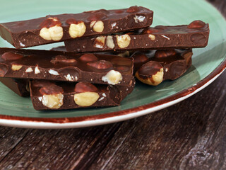 pieces of broken hazelnut chocolate on a mint green plate on wooden table, close up of dark chocolate withs nuts with copy space