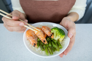 A girl in a white blouse and brown apron holds a plate of golden rice and three large king prawns in her hands. The dish is decorated with green beans and broccoli. Asian food concept