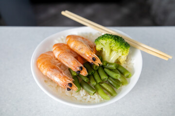 A bowl of rice, three king prawns, green beans and broccoli sits on a table in a dark kitchen. Nearby are chopsticks. Asian food concept