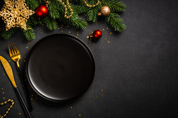 Christmas table with black plate, golden cutlery and holiday decorations. Top view on black.