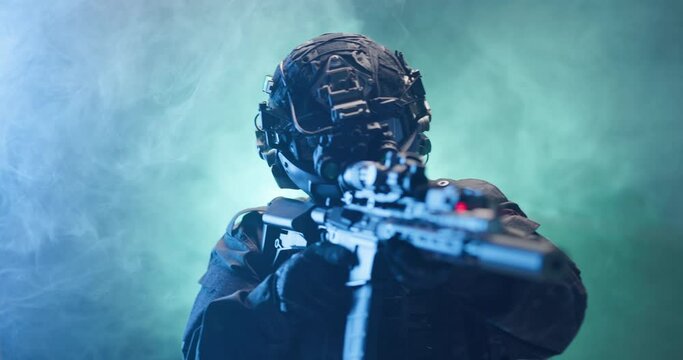Soldier fully equipped with tactical gear and protection aiming his rifle at enemy with blue and green smoke in the background.