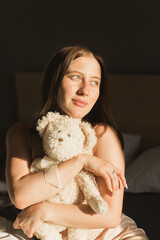 Portrait of girl is waking up in morning stretches in bed and sun shines from window. Happy young woman greets new sunny day and holds teddy bear toy