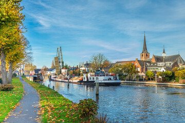 Cityscape of Koudekerk aan den Rijn with sailing sand loaded ship on the Oude Rijn river passes the opened drawbridge and on left bank the Brugkerk against blue sky with veil clouds