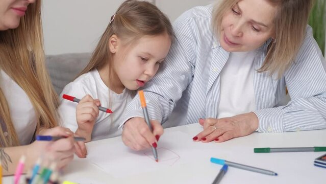 Creative family. Female relationship. Enjoying time. Happy little girl drawing with mother and grandmother together sitting desk in light room interior.