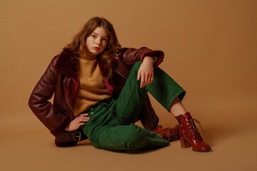 Fashionable freckled redhead girl wearing trendy autumn, winter outfit with green jeans, turtleneck sweater, aviator jacket, brown ankle boots, posing on beige background. Copy, empty space for text