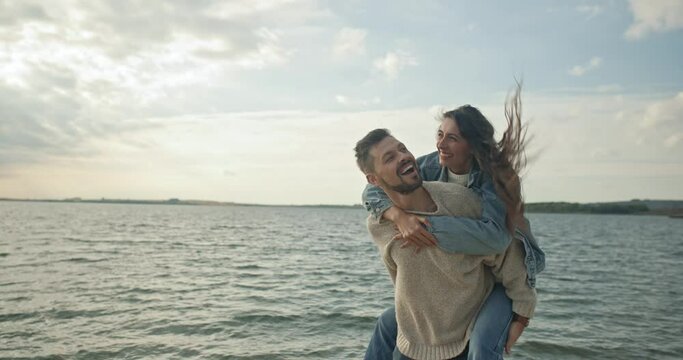 Cute lovely couple walking on beach near big beautiful lake. Attractive handsome man smiling carrying charming playful woman on back enjoying nature having date laughing having fun.