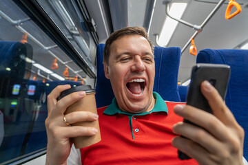 Overjoyed caucasian man looks at smartphone screen with mouth opened in train commuting to work. Excited man looking with wide open eyes into smartphone, holding paper coffee cup
