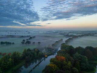 Aerial view of a rural landscape during sunrise in Belgium. Rural farm, corn fields, green fields and colorful fall foliage on the trees, sunlight and fog. Belgium, Europe. High quality photo