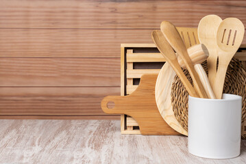 Wooden kitchen utensils, spoon, spatula, tongs, drainer and other utensils in a jar next to cutting boards and trivets, with a light wooden background. Copy space.