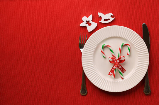 A white ribbed plate, two candy canes on it, a red festive ribbon with hearts, cutlery, white horse ans angel Christmas decor, red background, copy space