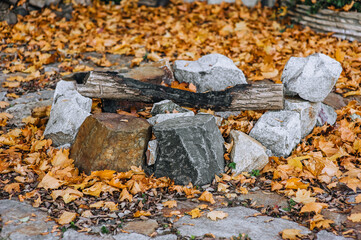 Homemade large fireplace made of stones and extinct firewood in nature in autumn with fallen yellow maple leaves. Photography of nature.
