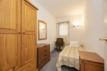 Fototapeta na wymiar A narrow bedroom with a youthful bed and mismatched wooden furniture and a varnished pine wardrobe
