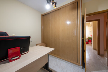 An office with a gaming chair, a designer PC and a built-in wardrobe with sliding oak doors and an access door made of the same material and stoneware floors.