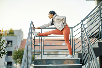A sportswoman is stretching her legs on railing while standing on stairs in urban exterior.
