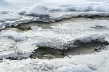 Shallow forest river covered with crystal snow and ice layer in winter, white patches over stones,...