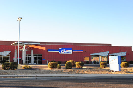Exterior view of a United States Post Office facility with red brick walls, official signage, front entrance, and parking lot, illustrating federal postal services and public infrastructure