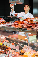 Interested young salesman of butcher store standing behind glass fridge showcase, weighing fresh sausage on scales .