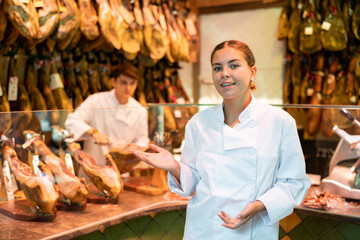 Cheerful young salesgirl in white uniform working in butcher shop, offering delectable Iberian jamon