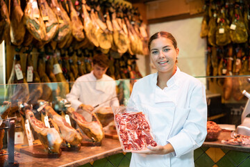 Portrait of positive young female butcher on background of rack with hanging various jamon in shop