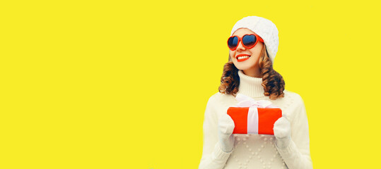 Winter portrait of happy smiling woman holding red gift box in her hands wearing white knitted hat, sweater on yellow background, blank copy space for advertising text