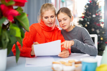 Smiling woman and her teenage daughter sitting at the table and writing wish list of Christmas...