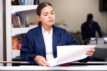 Attentive young female typographer in a blue uniform verifying printed sheet during work in the...