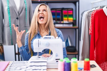 Blonde woman dressmaker designer using sew machine crazy and mad shouting and yelling with aggressive expression and arms raised. frustration concept.