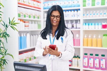 Middle age hispanic woman pharmacist using smartphone working at pharmacy