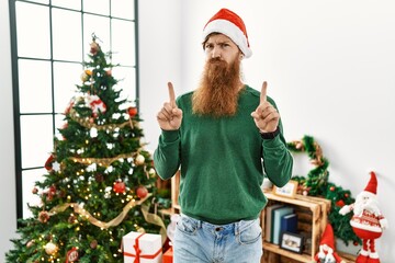Redhead man with long beard wearing christmas hat by christmas tree pointing up looking sad and...