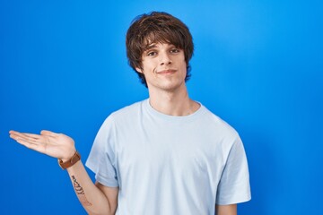 Hispanic young man standing over blue background smiling cheerful presenting and pointing with palm of hand looking at the camera.