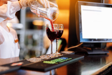 Bartender is pouring red wine into two long-stemmed wineglasses from a measuring cup on the bar counter. Blurred background. Party , cocktail, alcohol, beverage, restaurant, relax concept.