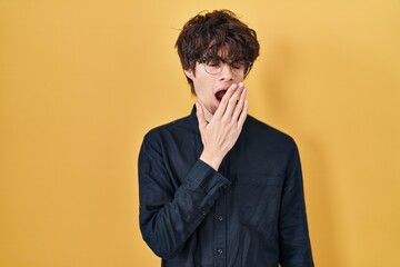 Young man wearing glasses over yellow background bored yawning tired covering mouth with hand. restless and sleepiness.