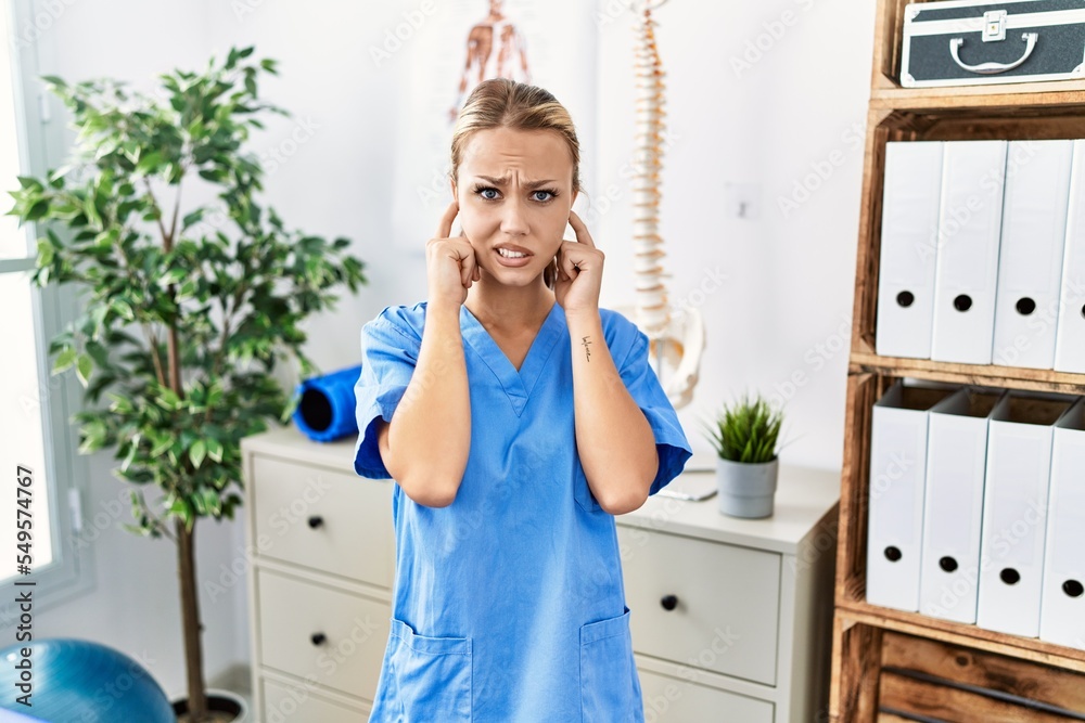 Poster young caucasian woman working at pain recovery clinic covering ears with fingers with annoyed expres