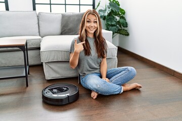 Young caucasian woman sitting at home by vacuum robot pointing finger to one self smiling happy and...