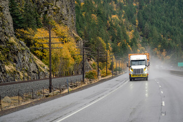 Fototapeta na wymiar White big rig industrial long haul semi truck transporting oversize load driving on the autumn road with rock mountain at raining weather