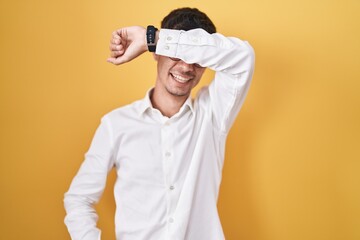 Young hispanic man standing over yellow background covering eyes with arm smiling cheerful and funny. blind concept.