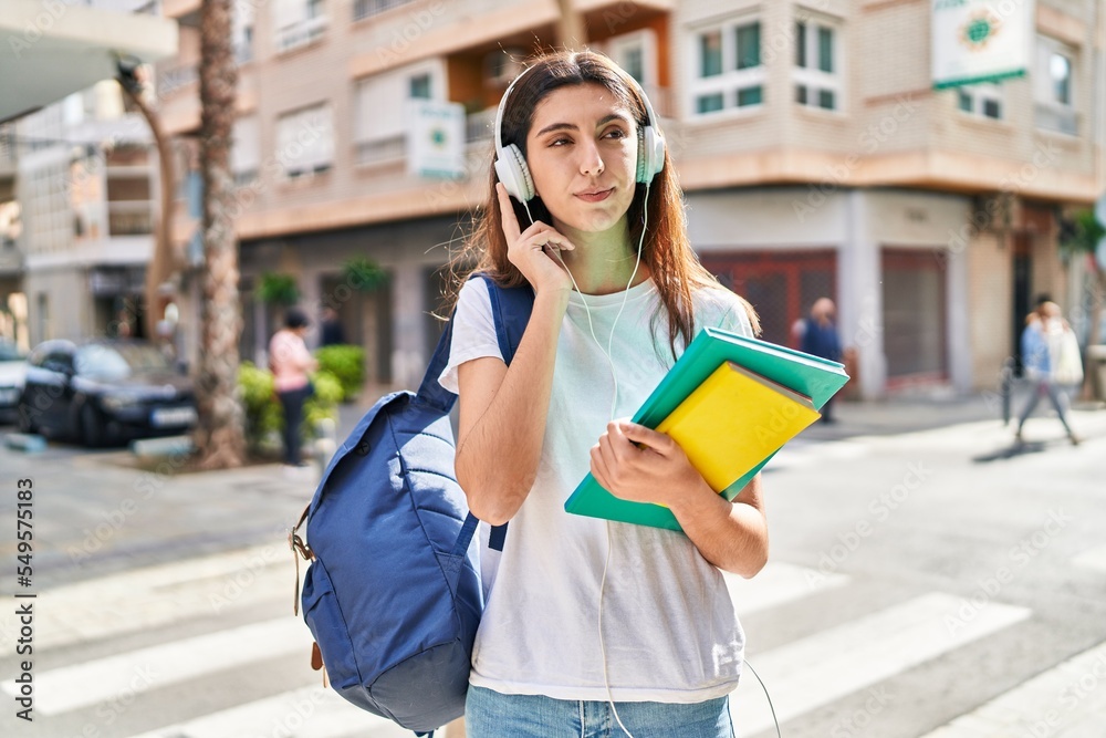 Wall mural Young beautiful hispanic woman student holding books using smartphone at street