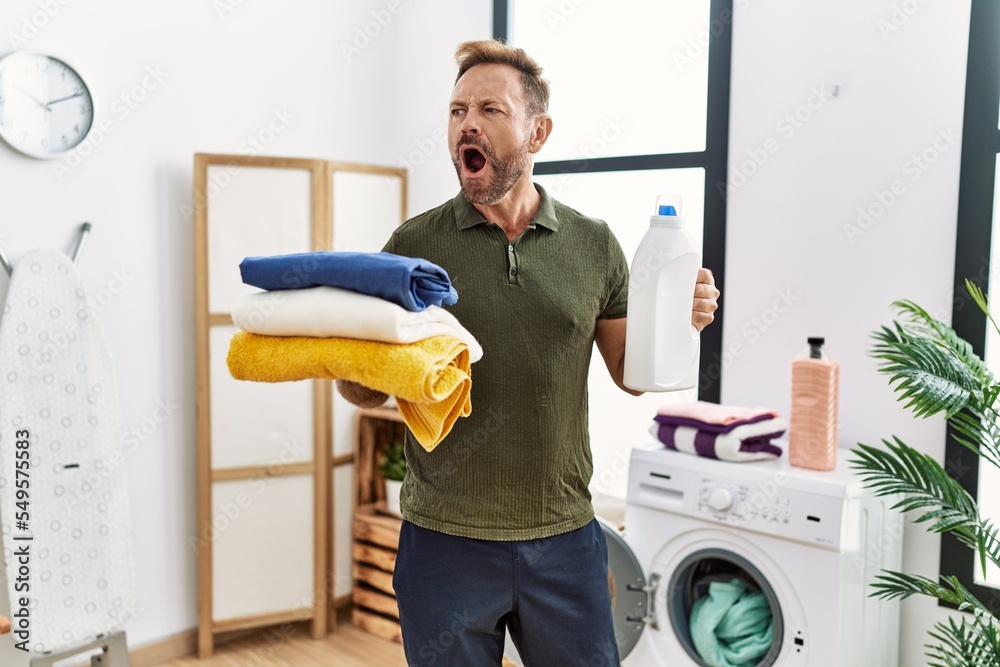 Sticker Middle age man holding clean laundry and detergent bottle angry and mad screaming frustrated and furious, shouting with anger. rage and aggressive concept.