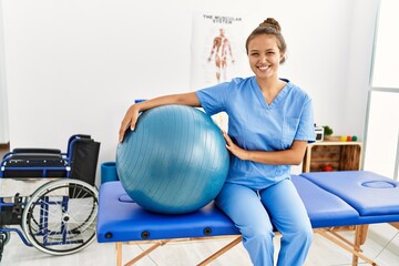 Young beautiful hispanic woman physiotherapist smiling confident holding fit ball at rehab clinic